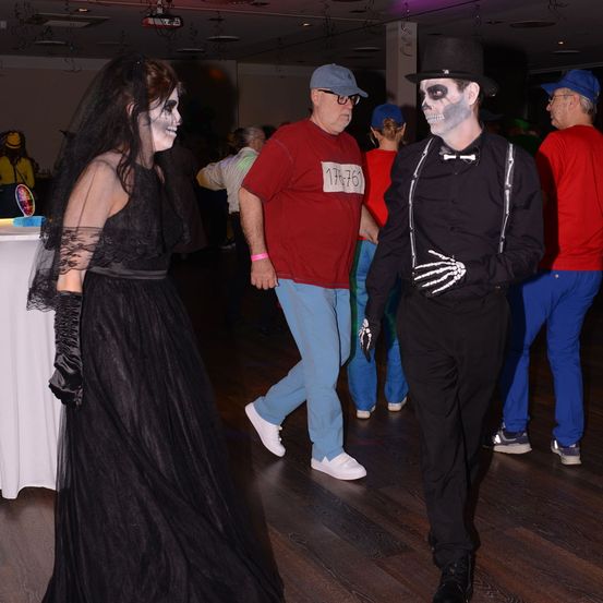 Eine Gruppe von Menschen ist in Kostümen bei einer Party. Eine Frau in einem schwarzen Kleid und Schleier und ein Mann in einem Skelettkostüm stehen nebeneinander.