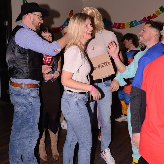 Eine Gruppe von Menschen tanzt bei einer Party. Eine Frau hält ein Schild mit der Aufschrift 'No-Filter'. Ein Mann in einem Cowboyhut hält eine Frau an der Taille. Hinter ihnen sind weitere Personen in bunten Kostümen gekleidet.