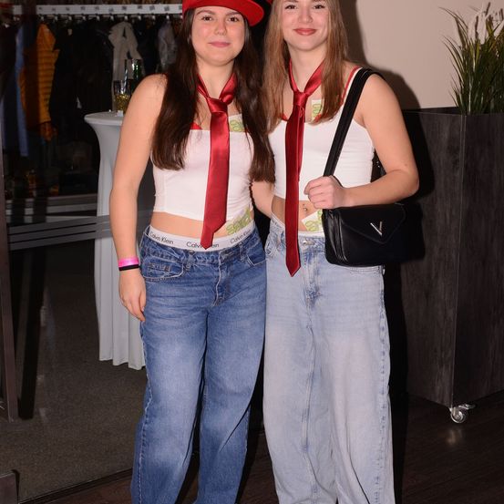Zwei Frauen stehen nebeneinander, beide tragen rote Krawatten, weiße Crop-Tops und Jeans. Die Frau auf der linken Seite trägt einen roten Hut.