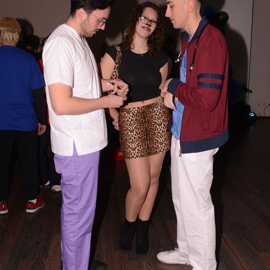 Drei Menschen stehen in einem Raum; ein Mann in einem weißen Mantel spricht mit einer Frau in einem Leopardenrock. Hinter ihnen steht ein Mann in einem roten Mantel und lauscht. Ein anderer Mann steht hinter ihnen. Der Raum ist schwach beleuchtet.