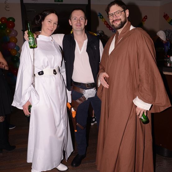 Drei Menschen in Kostümen stehen bei einer Veranstaltung zusammen und halten Bierflaschen in den Händen. Eine Frau in einem weißen Kleid und ein Mann in einem schwarzen West. Ein anderer Mann trägt einen braunen Umhang.