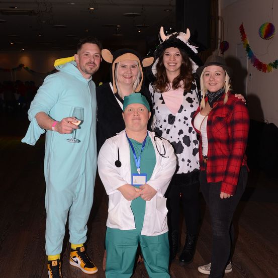Fünf Menschen in Kostümen stehen lächelnd zusammen. Ein Mann in einem hellblauen Kostüm, eine Frau in einem Kuhkostüm und eine andere Frau in einer roten karierten Jacke. Ein Mann in einem Arztumhang und eine Frau in einem Mauskostüm. Sie sind in einem Raum mit Holzboden und Ballons an der Wand.
