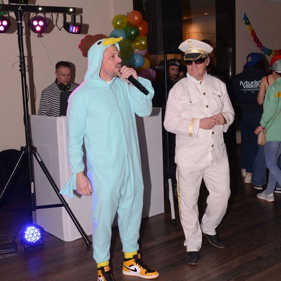 Ein Mann in einem blauen Kostüm mit Vogelkopfbedeckung hält ein Mikrofon. Ein Mann in einem weißen Uniform steht neben ihm. Sie befinden sich auf einer Tanzfläche mit einem DJ-Pult.
