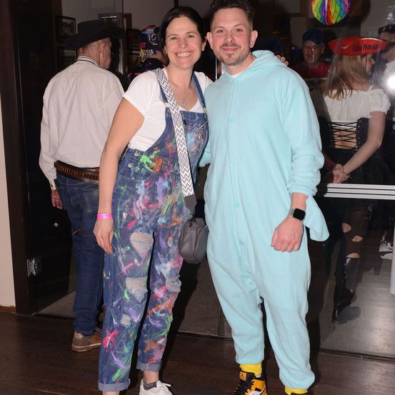 Ein Paar in bunter Kleidung, die Frau in einem besprühten Jumpsuit, der Mann in einem hellblauen Overall, beide lächeln, mit anderen Menschen im Hintergrund.