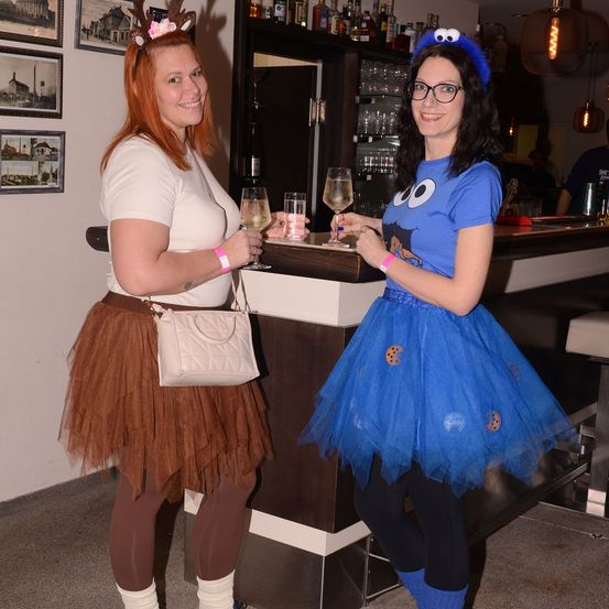 Zwei Frauen in Kostümen, eine mit Rentiergeweih, die andere als Cookie Monster verkleidet, halten Weinflöten in einer Bar.