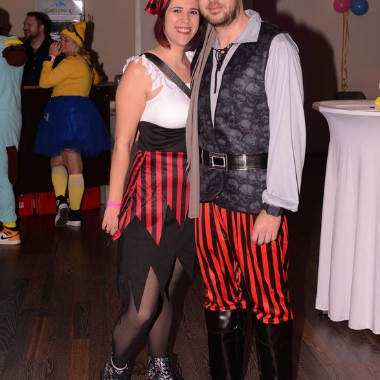 Ein Mann und eine Frau in Piratenkostümen lächeln für ein Foto, mit anderen in verschiedenen Kostümen und Dekorationen im Hintergrund.