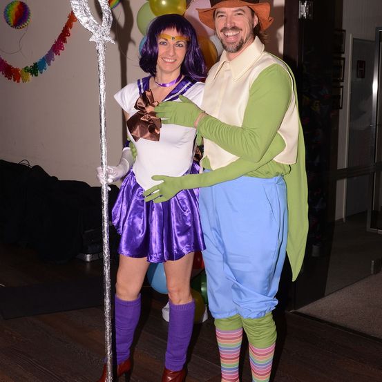 Ein Mann und eine Frau posieren in einem Raum zusammen, in Kostümen gekleidet. Der Mann trägt ein grünes Kostüm und die Frau ist als Matrose gekleidet. Sie stehen auf einem Holzboden, mit Luftballons und Dekorationen im Hintergrund.