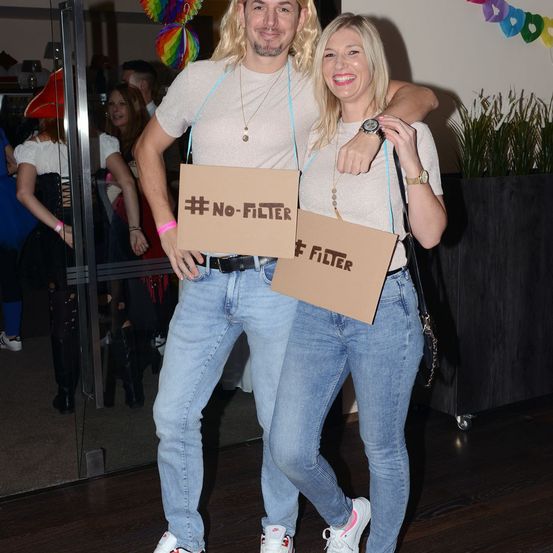 Ein Mann und eine Frau in Kostümen posieren mit Schildern, die #No-Filter und #Filter anzeigen. Sie sind bei einer Veranstaltung, wahrscheinlich einer Party, mit anderen in Kostümen hinter ihnen.
