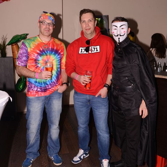 Drei Männer auf einer Party, einer in einem Tie-Dye-Shirt, einer in einer roten Kapuzenjacke mit 17671 und einer in einer Maske. Sie stehen eng beieinander und halten Getränke in den Händen.