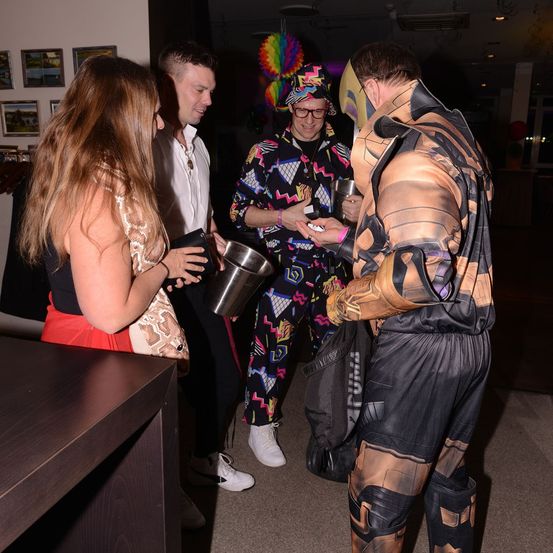 Mehrere Menschen stehen und unterhalten sich in einem Raum, eine Person in Kostüm, eine andere mit einem silbernen Eimer, und eine Frau mit einer Handtasche.