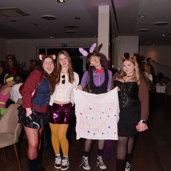 Fünf Frauen in Kostümen posieren für ein Foto, eine hält ein Schild mit der Aufschrift 'Lasst uns feiern'. Sie stehen in einem Raum mit Holzböden und Stühlen. Andere Leute, ebenfalls in Kostümen, sind im Hintergrund.