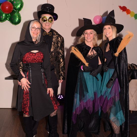 Eine Gruppe von vier Personen in Kostümen steht in einem Raum, zwei halten Besen in den Händen. Die Frau auf der linken Seite trägt einen schwarzen Umhang. Hinter ihnen hängen Luftballons an der Wand.