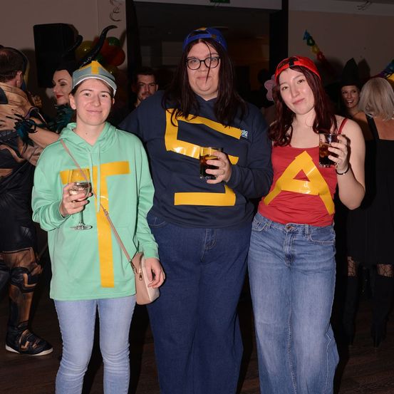 Drei Frauen in Kostümen halten Getränke, eine trägt ein 'S'-Shirt, eine andere 'A' auf einem roten Shirt. Ein Mann mit einem gehörnten Hut steht hinter ihnen.