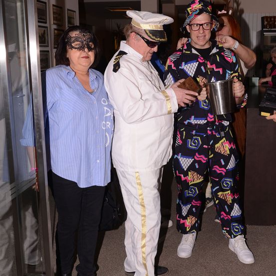 Drei Menschen in Kostümen stehen. Eine Frau mit einer Maske, ein Mann in einem Matrosenanzug und ein Mann in einem bunten Anzug, der einen Eimer hält.