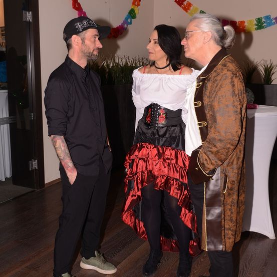 Ein Mann in einem schwarzen Shirt und Hut steht mit einer Frau in einem Piratenkostüm und einer älteren Frau in einem braunen Mantel. Sie scheinen sich bei einer Veranstaltung zu befinden.