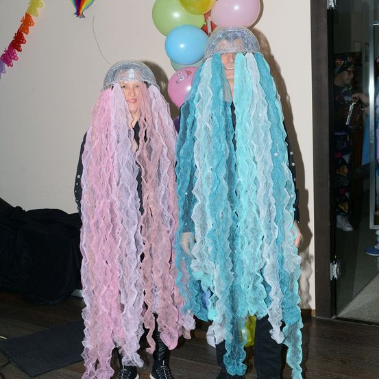 Zwei Menschen in rosa und blauen Tentakelkostümen, die vor einem Hintergrund mit bunten Luftballons stehen.