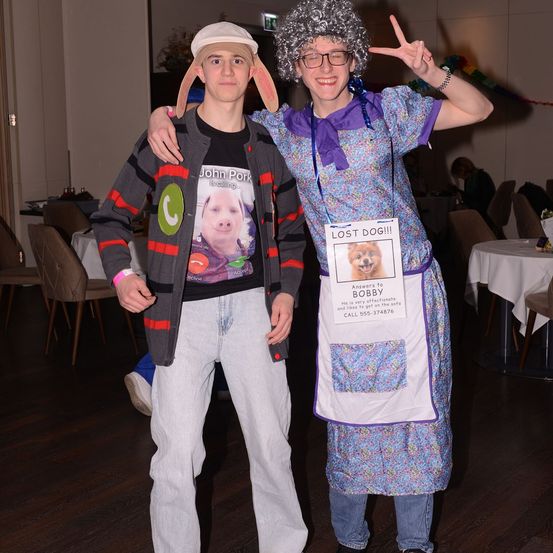 Zwei Menschen in Kostümen stehen in einem Raum. Einer trägt ein Schweine-Kostüm und der andere eine Blumenschürze mit einem Plakat für einen vermissten Hund. Sie posieren für ein Foto.
