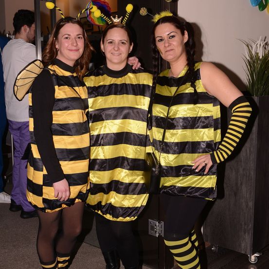 Drei Frauen in Bienenkostümen posieren für ein Foto. Eine von ihnen hat eine Krone auf dem Kopf. Hinter ihnen ist ein Mann in einem weißen T-Shirt und lila Hose zu sehen.