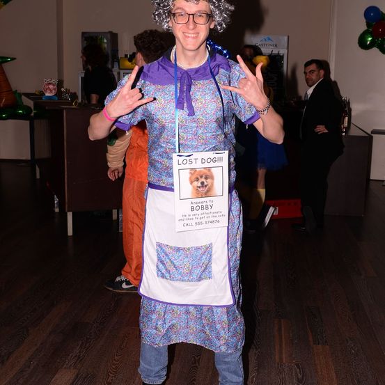 Eine Frau mit Perücke und Brille, gekleidet in einer Blumenschürze, hält ein Schild mit einem vermissten Hund namens Bobby und steht in einem Raum mit Luftballons und anderen Leuten.
