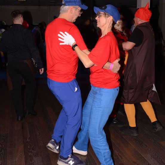 Zwei Menschen in roten T-Shirts, blauen Hosen und Mützen tanzen in einem Raum. Ein Mensch mit rotem Hut steht hinter ihnen. Ein anderer Mensch in schwarzer Kleidung steht in der Nähe.