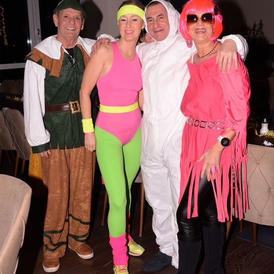 Vier Menschen in bunten Kostümen posieren für ein Foto. Ein Mann in Grün steht neben einer Frau in Pink und Grün. Ein Mann in Weiß steht neben ihnen. Eine Frau in Pink und Schwarz steht rechts.