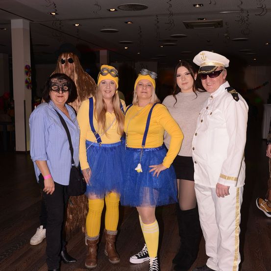 Fünf Personen in Kostümen posieren für ein Foto. Zwei sind in Minion-Kostümen, einer ist als Pirat verkleidet und zwei sind in normaler Kleidung. Sie sind drinnen in einem schwach beleuchteten Raum.