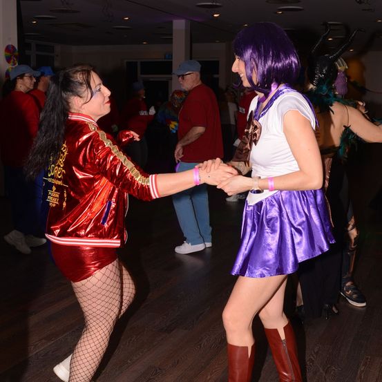 Zwei Frauen tanzen in einem Club, eine in einem roten Kostüm und die andere in Lila. Sie halten Händchen, während andere um sie herum tanzen.