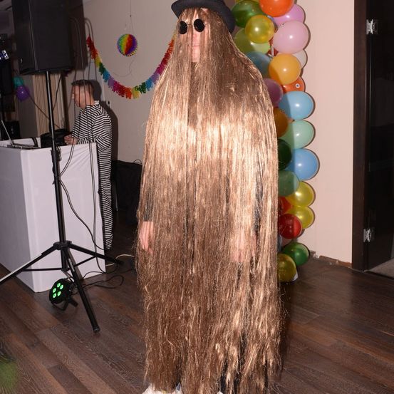 Eine Person mit langen goldenen Haaren steht vor einer bunten Ballonwand. Eine andere Person steht hinter einem Tisch mit Laptop und Lautsprecher.