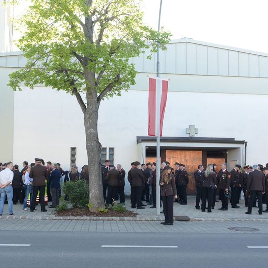Eine Gruppe uniformierter Personen steht vor einem Gebäude mit einem Fahnenmast und einem Baum. Sie warten in einer Schlange, einige halten Hüte.