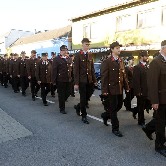 Eine Militärparade von uniformierten Personen, die in einer Reihe marschieren und vor Gebäuden und einem geparkten Auto vorbeiziehen.