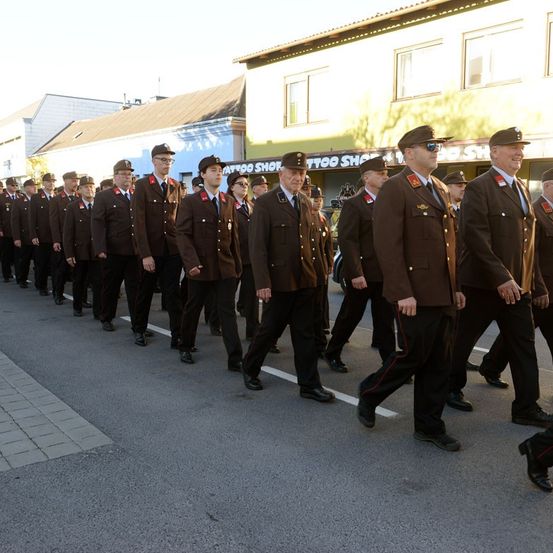 Eine Gruppe uniformierter Personen, wahrscheinlich eine militärische oder polizeiliche Einheit, marschiert die Straße entlang. Im Hintergrund befinden sich Gebäude und ein Geschäft.