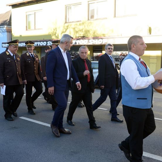 Eine Gruppe formell gekleideter Männer, darunter zwei in Militäruniformen, gehen in einer Reihe auf einer Straße. Hinter ihnen befindet sich ein Gebäude mit einem Schild, das Tätowierungsstudio lautet.