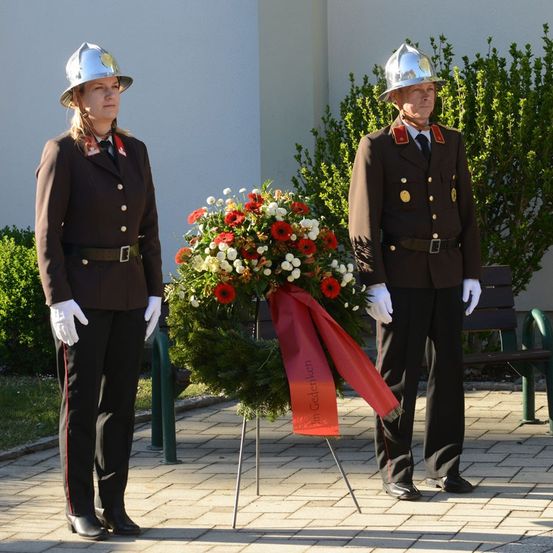 Zwei Feuerwehrleute in Uniform stehen neben einer Schleife und einem roten Band. Sie tragen weiße Handschuhe und stehen in einem Garten.
