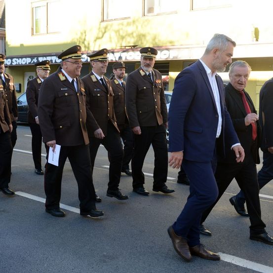 Eine Gruppe uniformierter Offiziere in Militäruniformen geht vor zwei Männern in Anzügen, einer von ihnen hält ein Papier. Sie sind auf einer Straße, mit Gebäuden und geparkten Autos im Hintergrund.