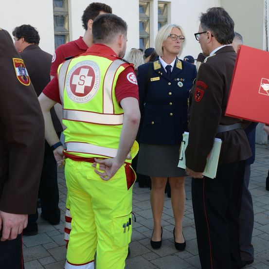 Eine Frau in Uniform steht zwischen zwei Männern, beide in Militäruniformen, draußen in der Nähe eines Gebäudes. Der Mann auf der linken Seite trägt eine reflektierende Weste mit einem Abzeichen.