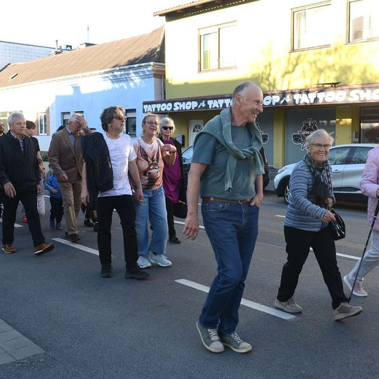 Mehrere Menschen gehen über eine Straße, ein Mann vorn trägt einen Schal. Dahinter eine Frau mit einem Stock und ein geparktes Auto. Gebäude säumen die Straße.