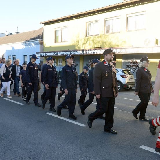 Eine Gruppe von Menschen in Uniformen und Hüten marschiert vor einem Tattoo-Laden die Straße entlang.