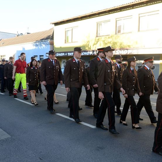 Eine Gruppe von Personen in Uniformen marschiert in einer Parade die Straße hinunter. Die Uniformen sind braun mit rotem Besatz. Ein Mann in einem roten Shirt und neongelben Hosen ist vorne.