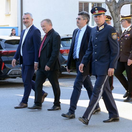 Eine Gruppe von Männern in formeller Kleidung geht, wahrscheinlich bei einem formellen Anlass. Sie werden von Personen in Militäruniformen begleitet.