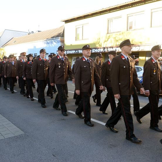 Eine Reihe von Offizieren in braunen Uniformen mit roten Streifen marschiert die Straße hinunter und passiert ein Gebäude mit einem Ladenschild.