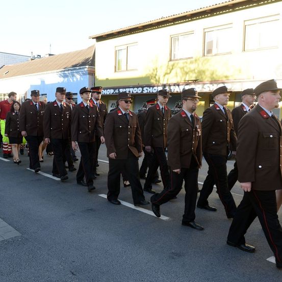 Eine Gruppe uniformierter Männer marschiert auf einer Straße, vermutlich bei einer Parade, vor einem gelben Gebäude.