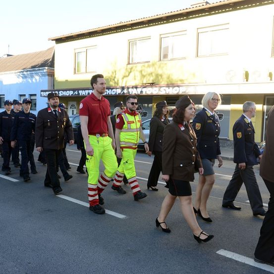 Eine Gruppe von Menschen in Uniformen, die an einer Parade auf der Straße teilnehmen. Der Mann in der Mitte trägt reflektierende gelbe Hosen.