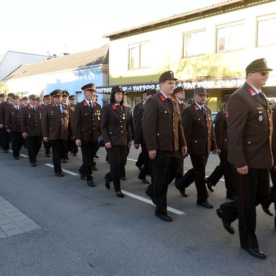 Eine Gruppe uniformierter Personen marschiert in Formation die Straße hinunter, trägt Medaillen und Mützen. Hinter ihnen befindet sich ein Gebäude mit einem Tattoo-Shop-Schild.