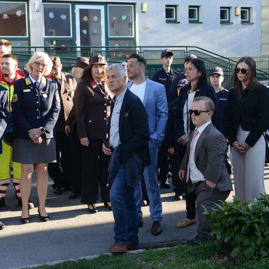 Eine Gruppe von Menschen, darunter uniformierte Personen und andere, steht vor einem Gebäude. Einige tragen Sonnenbrillen und formelle Kleidung.