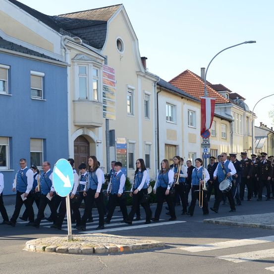Eine Blaskapelle mit Instrumenten und Uniformen läuft auf der Straße vor Gebäuden. Ein blauer Fahnenmast mit Flagge steht an der Seite.