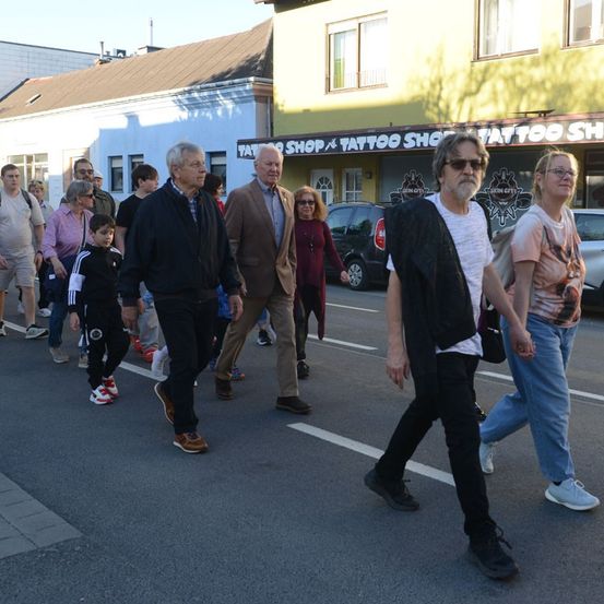 Eine Gruppe von Menschen geht eine Straße entlang, vermutlich auf einer Tour. Zwei Männer vorne führen die Gruppe an. Dahinter befindet sich ein Tattoo-Shop mit geparkten Autos.