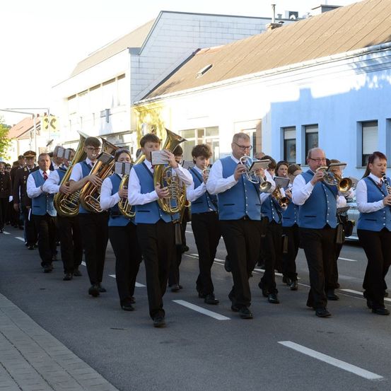 Eine Marschkapelle in blauen Westen und schwarzen Hosen spielt verschiedene Blechblasinstrumente, darunter Tubas, Trompeten und Posaunen, während sie in Formation auf einer Straße geht. Gebäude säumen die Straße, und die Schatten der Gebäude fallen auf den Boden.