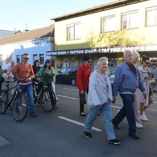 Eine Gruppe von Menschen, darunter ältere Erwachsene, geht vor einem Tattoo-Laden auf der Straße, einige fahren Fahrrad.