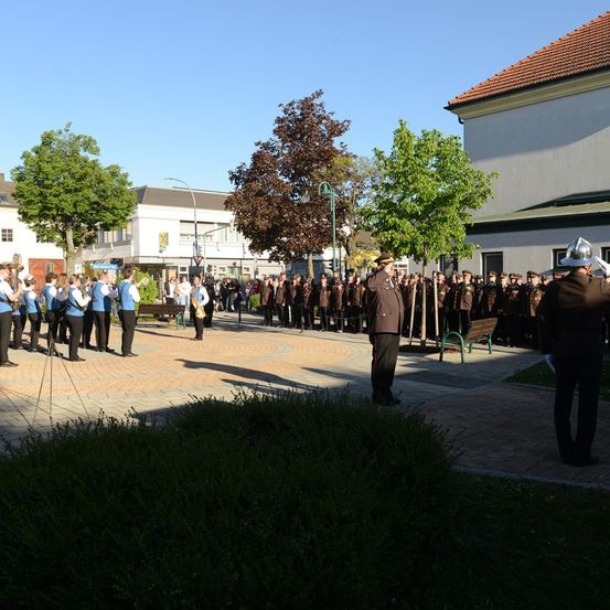 Eine Gruppe von Personen in Uniformen steht in einem Außenbereich. Sie scheinen eine Parade durchzuführen. Eine Bank und Bäume befinden sich hinter ihnen.