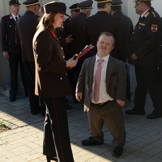 Eine Frau in Uniform steht vor einem Mann in Anzug, beide halten Gegenstände. Dahinter stehen mehrere uniformierte Beamte in einer Reihe.